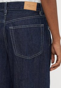 Tumši zili džinsa džinsi ar kontrastējošu baltu šuvju apdari. Ir aizmugurējā kabata un gaiši brūna ādas etiķete ar "perfect jeans" zīmolu.