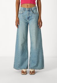 Svetlomodré, široké džiny vyrobené z denimu s vysokým pásom, bočnými vreckami a viditeľnými stehovými detailmi. Nosené so svetlými sandálmi.