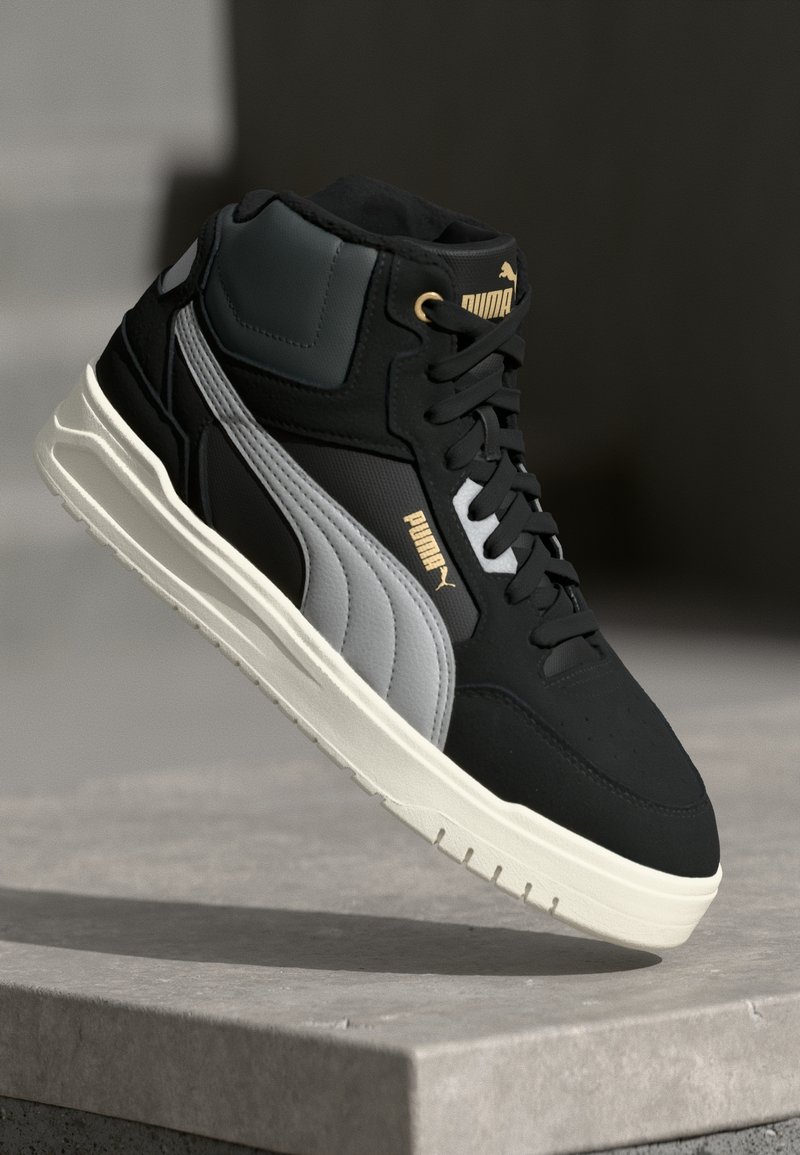 Baskets montantes Puma noires et grises avec semelle blanche, accents dorés sur le logo et lacets, présentées flottant au-dessus d'une surface en béton.