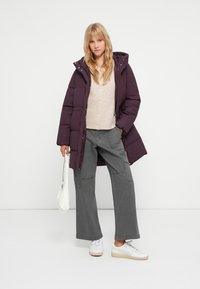 Ullsfylt lilla parkdress med hette, beige cardigan, løse grå cargo-bukser og hvite sneakers. Modellen holder en hvit veske foran en ensfarget bakgrunn.