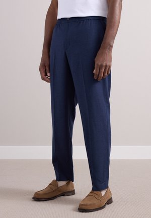 Homme portant un pantalon bleu marine sur mesure, des chaussettes blanches, des mocassins en daim marron et une chemise blanche, debout sur un sol recouvert d'un tapis beige.