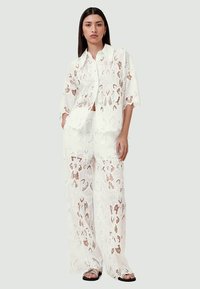 Ensemble deux pièces en dentelle blanche comprenant une chemise à boutons à manches courtes et un pantalon large, mettant en avant des motifs floraux et une texture légère.