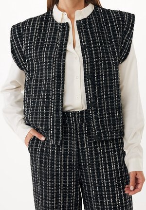 Vrouw draagt een zwart-wit geruit vest en bijpassende broek over een witte blouse met knopen, met één hand in de zak.