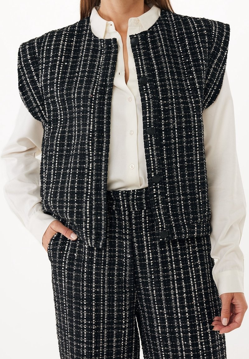 Femme portant un gilet à carreaux noirs et blancs et un pantalon assorti par-dessus un chemisier blanc boutonné, avec une main dans la poche.
