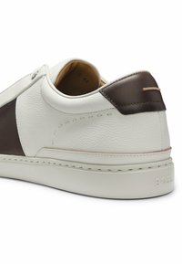 Baskets blanches en cuir avec patch marron au talon, dessus texturé et semelle épaisse montrant partiellement la pointure à l'intérieur.