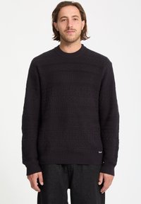 Sort striktrøje med teksturerede mønstre, ribbede manchetter og rund halsudskæring. Båret med mørke jeans, der viser en afslappet pasform.