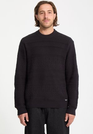 Suéter negro de punto con patrones texturizados, puños acanalados y un escote redondo. Se lleva con jeans oscuros, mostrando un ajuste relajado.