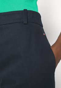 Marineblauwe broek met een gladde textuur, voorzien van een riemlus en een klein geborduurd logo in rood en wit aan de zijkant.