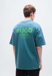 Τυρκουάζ υπερμεγέθη T-shirt με έντονο πράσινο γραφικό κείμενο στην πλάτη, που περιλαμβάνει τις λέξεις "HUGO" και "MCMXCIII", κατασκευασμένο από μαλακό βαμβάκι.