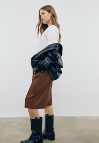 Pantaloni corti marroni su misura, t-shirt bianca, giacca di pelle nera appesa sulla spalla e stivali neri al ginocchio con dettagli a fibbia.