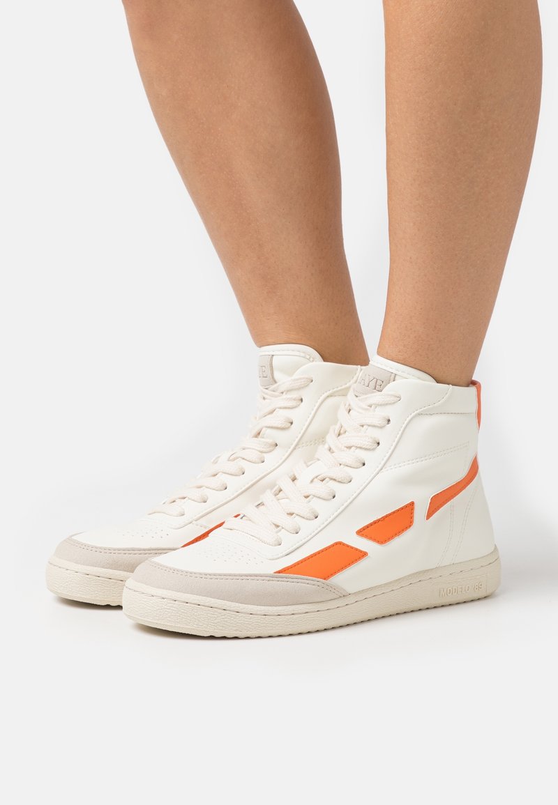 Baskets montantes blanches avec accents orange portées sur jambes nues, dotées d'un design à lacets et d'une semelle beige clair.