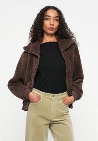Bruine fleece zip-up jas met hoge kraag, gecombineerd met een zwarte top en lichtgroene corduroy broek. De jas heeft ribgebreide manchetten.