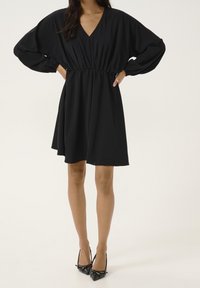 Robe noire avec un décolleté en V profond, taille cintrée et manches amples et bouffantes. Associée à des chaussures à talons hauts noires ornées de nœuds.