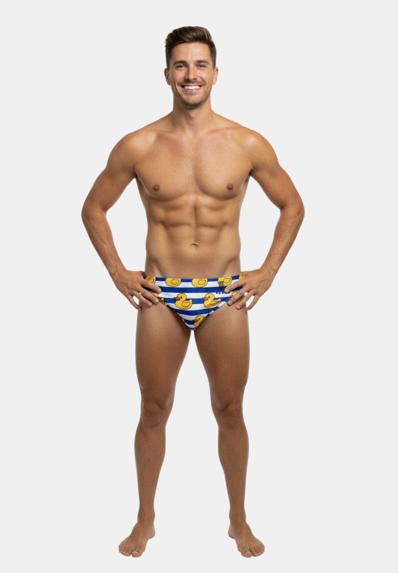 Hombre sin camisa y sonriendo, de pie con las manos en las caderas, vistiendo un bañador de rayas azules y blancas con patrones de patitos de goma amarillos.