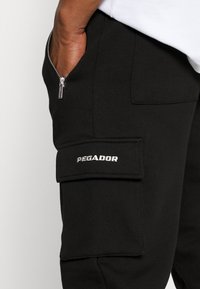 Svarta cargobyxor med en dragkedjeficka, som har en silverfärgad "PEGADOR"-logotyp och en texturerad tyg. Synliga sömmar och en strukturerad design.