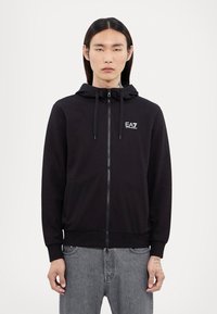 Svart zip-up hoodie med justerbar dragsko på huvan, fickor fram, ribbade muddar och en liten logo på bröstet. Klassisk och avslappnad design.