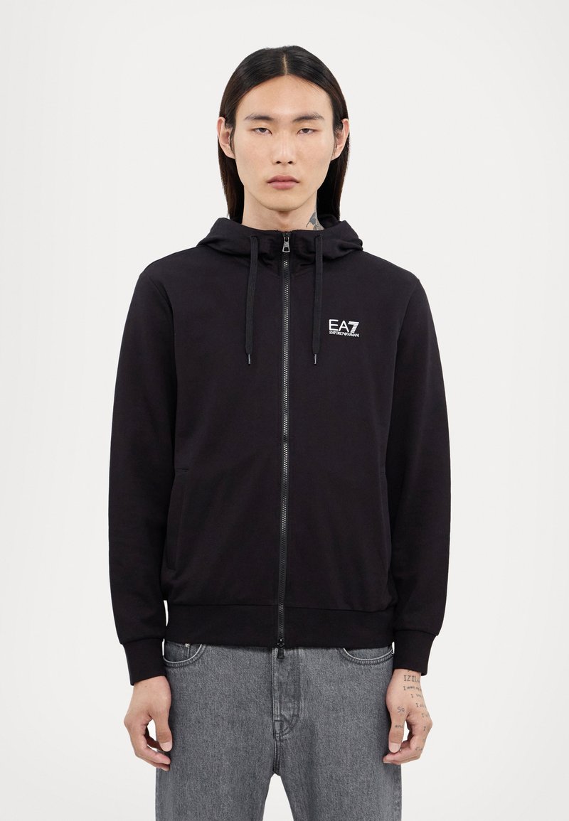 Svart zip-up hoodie med justerbar dragsko på huvan, fickor fram, ribbade muddar och en liten logo på bröstet. Klassisk och avslappnad design.