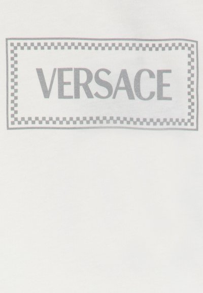 Balta T-kreklā ir pelēks taisnstūrveida logotips ar vārdu "VERSACE" treknrakstā, ko ieskauj šaha raksts.