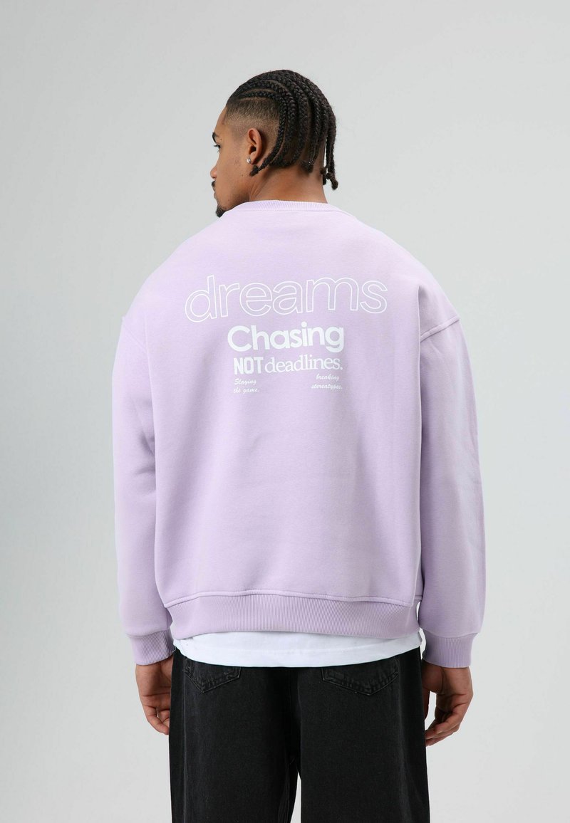 Sweat-shirt lavande à coupe ample ; comporte un texte imprimé blanc au dos : "dreams Chasing NOT deadlines." Bords-côtes aux poignets et à l'ourlet.