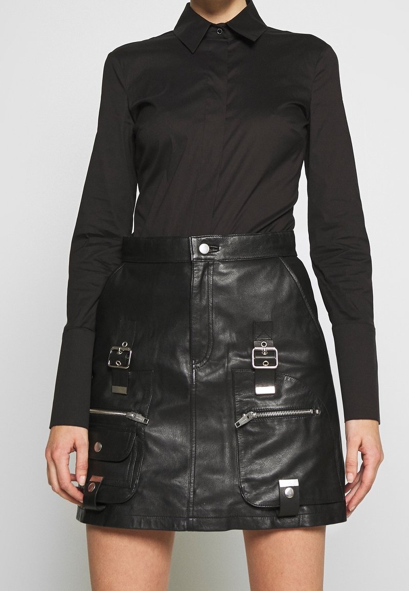 Gonna nera in pelle con vita alta, tasche con zip frontali e accenti metallici. Abbinata a una camicia nera a maniche lunghe con colletto.