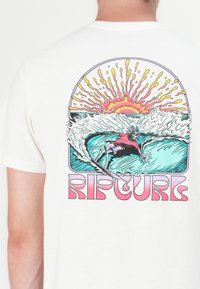 Hvit bomullst-skjorte med et fargerikt grafisk trykk av et skjelett som surfer med bølger og en sol, merket "RIPCURL" med fet pink skrift.