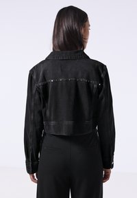 Lyhennetty musta farkkutakki, jossa on kaulus, napitus ja koristeellisia niittejä rivissä takana. Sileä pinta, kiinteä malli.