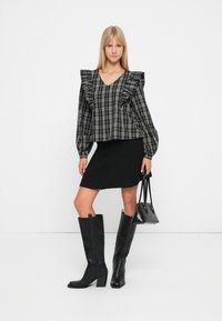 Haut à carreaux noir et gris avec des épaules à volants, associé à une jupe en tricot noire et des bottes noires à talons jusqu'aux genoux, tenant un petit sac noir.