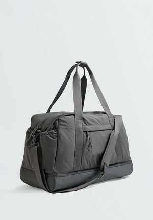Sac polochon gris foncé avec doubles poignées, bandoulière réglable, poche latérale zippée et fond renforcé sur un fond clair uni.
