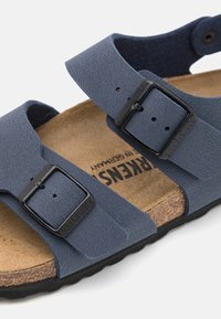 Blå, teksturert syntetisk sandale med to justerbare svarte spenner, kork fotbed og svart gummisåle. Lett og åpen tå design.