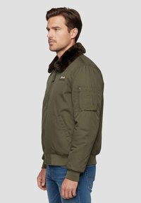 Olijfgroene bomberjack met een bruine bontkraag, ritssluiting en zijzakken. Heeft een patch op de borst en details op de mouwzak.