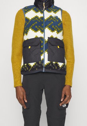 Personne portant un gilet en polaire à motifs avec des poches noires, des manches tricotées jaune moutarde et un pantalon cargo noir avec un logo patch.
