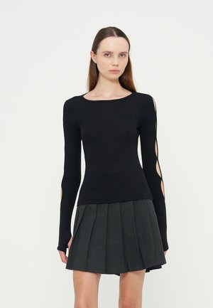 Jeune femme aux longs cheveux bruns porte un haut noir à manches longues avec découpes et une jupe plissée foncée, debout devant un fond uni.