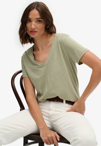 Lichtgroen T-shirt met korte mouwen en V-hals, met een zachte textuur en een subtiel gestreept patroon, gedragen met witte aansluitende broek en een bruine gevlochten riem.