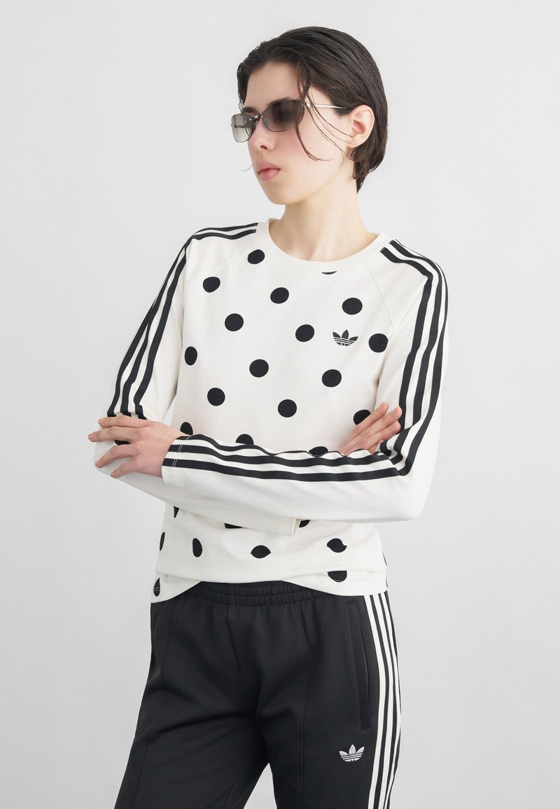 Isik, kes kannab musti päikeseprille, valget pusat mustade täppide ja triipudega ning musti pükse valgete triipudega, käed rüpes.