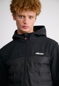 Černá kapucňová bunda se zipem, s prošívanou spodní částí, matným povrchem a logem "ellesse" na hrudi.