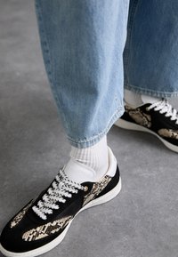 Baskets noires et crème avec un motif imprimé serpent, des lacets blancs et des accessoires dorés, associées à un jean bleu clair et des chaussettes blanches.