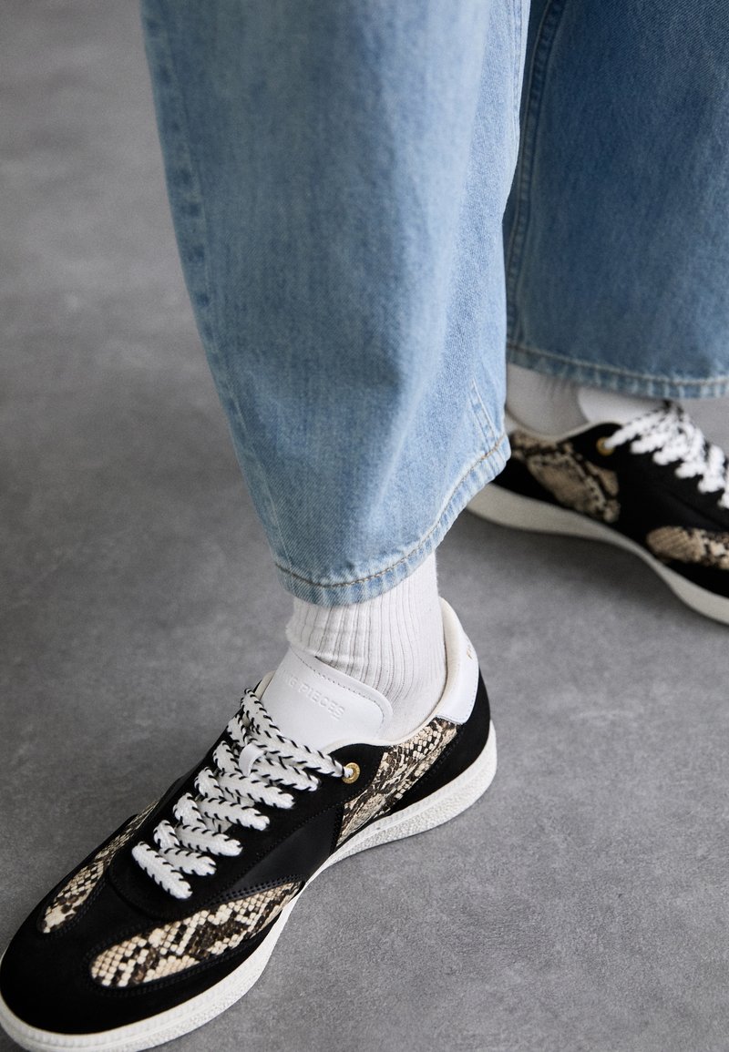 Baskets noires et crème avec un motif imprimé serpent, des lacets blancs et des accessoires dorés, associées à un jean bleu clair et des chaussettes blanches.