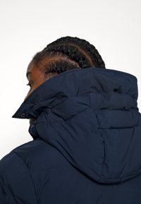 Granatowa kurtka typu puffer z wysokim kołnierzem i widocznymi przeszyciami, o gładkiej, matowej fakturze i zamkiem błyskawicznym z przodu.