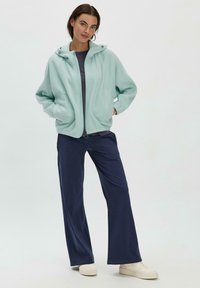 Helle mintfarbene Fleecejacke mit Kapuze, Reißverschluss und Seitentaschen, kombiniert mit dunkelblauen, weit geschnittenen Hosen und beigen Sneakers.