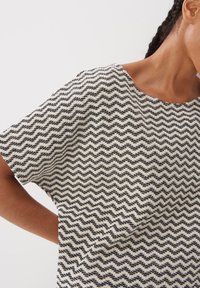 T-shirt à manches courtes avec une base crème et un motif zigzag noir, un col rond, et une coupe décontractée fabriquée en tissu texturé.
