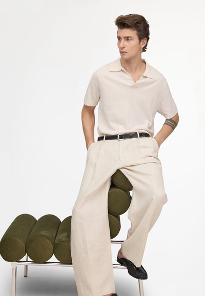 Giovane uomo con capelli corti e tatuaggio sul braccio, indossa una polo beige e pantaloni a gamba larga, seduto di lato su una sedia moderna verde oliva.