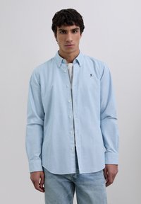 Chemise à rayures bleues et blanches avec boutons, en coton, dotée de manches longues, d'un col et d'un petit logo sur la poitrine. Portée par-dessus un T-shirt gris clair.