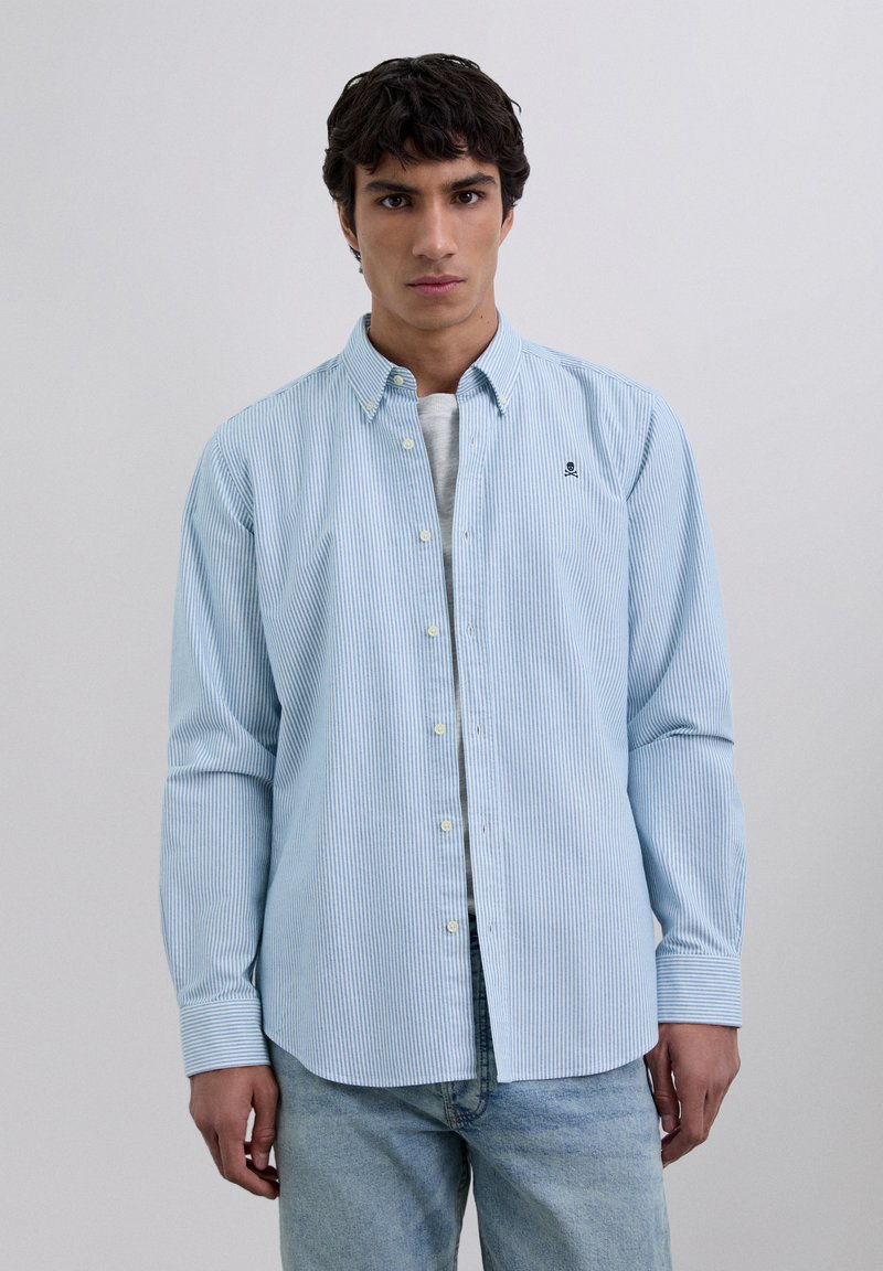 Chemise à rayures bleues et blanches avec boutons, en coton, dotée de manches longues, d'un col et d'un petit logo sur la poitrine. Portée par-dessus un T-shirt gris clair.