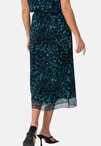 Robe longue couleur sarcelle avec un motif léopard. Fabriquée en tissu léger avec un ourlet transparent. Portée avec des sandales à talons noires.