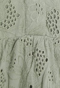 Ljust grönt tyg med intrikat broderi som visar blommiga och geometriska mönster, inklusive utskärningar och upphöjda texturer.