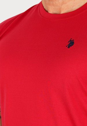 Piros, kerek nyakú póló, bal oldalon egy kis sötét hímzett polo játékos logóval, amelyet egy személy visel.