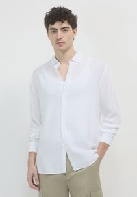 Giovane uomo con capelli ricci, indossa una camicia bianca con bottoni sopra una maglietta bianca e pantaloni beige, sta in piedi con una mano in tasca.