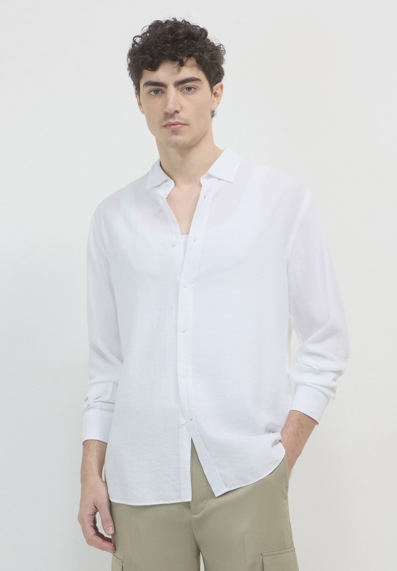 Giovane uomo con capelli ricci, indossa una camicia bianca con bottoni sopra una maglietta bianca e pantaloni beige, sta in piedi con una mano in tasca.