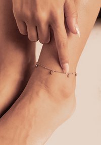 Cavigliera in oro rosa con piccoli charm circolari, caratterizzata da un design di catena delicato. La tonalità della pelle è visibile sul piede e sulla caviglia, mettendo in risalto i dettagli della gioielleria.