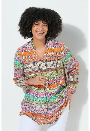 Smilende kvinde med krøllet hår iført en farverig, mønstret tunika med orange smileys og blomsterdesigns, stående mod en hvid baggrund.